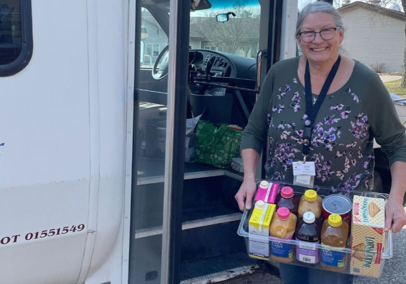 PHOTO 1 Public Health An older woman stands in front of a mini-bus holding a bin of snacks and juices.
