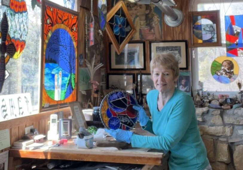 PHOTO-1-Remarkable-artists-open-their-Houston-County-studios-for-regional-art-tour-by-Rose-Korabek-April-2025 A woman in a turquoise shirt holds a piece of stained glass while she sits at a table surrounded by other pieces of art.