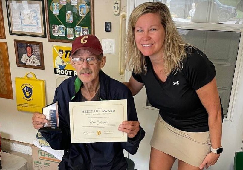Inside a room filled with framed sports memorabilia, an older man wearing glasses and a maroon baseball cap sits facing the camera while holding a framed certificate and a small glass award. The certificate reads “Heritage Award.” A woman with shoulder-length wavy blond hair stands beside him, leaning in and smiling toward the camera with one hand resting near his shoulder. Behind them are framed photos, vintage signs, and a window, creating a warm, personal setting that reflects community history and recognition.
