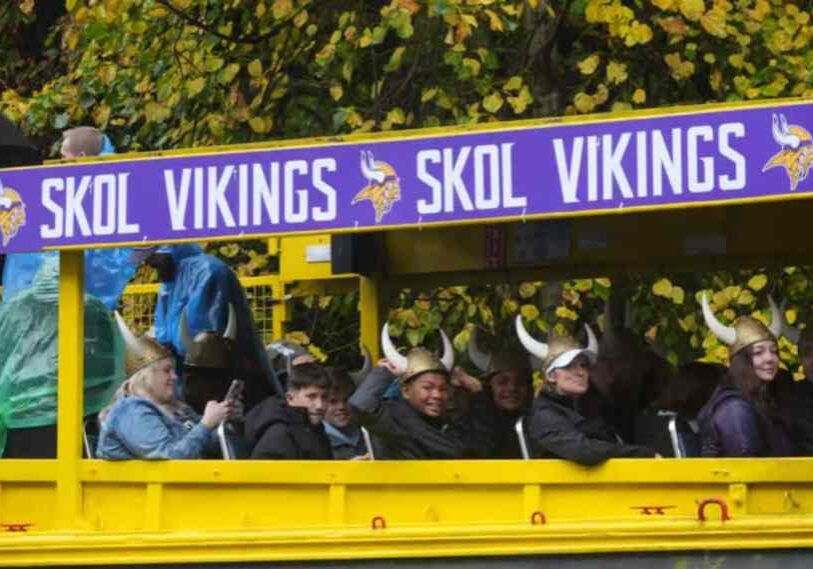 PHOTO 1 SKOL Vikings A group of Minnesota Vikings fans ride a bright yellow open-air bus decorated with large purple signs reading “SKOL VIKINGS” and the team’s horned-Viking logo. The fans, wearing gold Viking helmets with white horns, smile and wave despite the rainy weather. Some wear ponchos or hold umbrellas, and autumn trees with orange and green leaves fill the background.