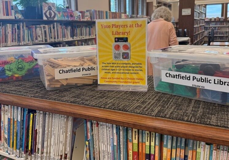 Wide view inside the Chatfield Public Library showing three clear plastic STEAM activity bins displayed on a long bookshelf table. One bin contains colorful craft and sensory materials, another holds wooden blocks, and a third contains building toys and supplies. A bright yellow sign in the center promotes Yoto Players available at the library for children’s educational audio stories and music. Rows of children’s books fill the shelves below and behind the display. Large windows along the left side bring natural light into the modern library space while a patron browses books in the background.