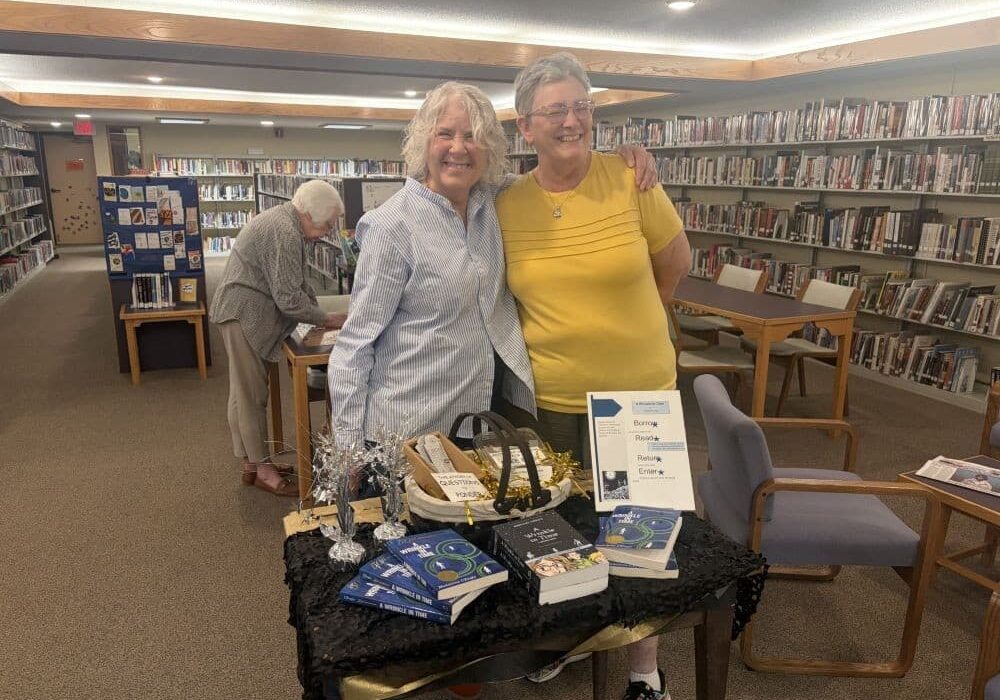 Two women stand side by side smiling in a library, arms around each other, behind a small display table covered with books and decorative items. The table includes stacked books, a basket, and a small sign. Bookshelves filled with rows of books line the background, and another person browses at a table behind them.
