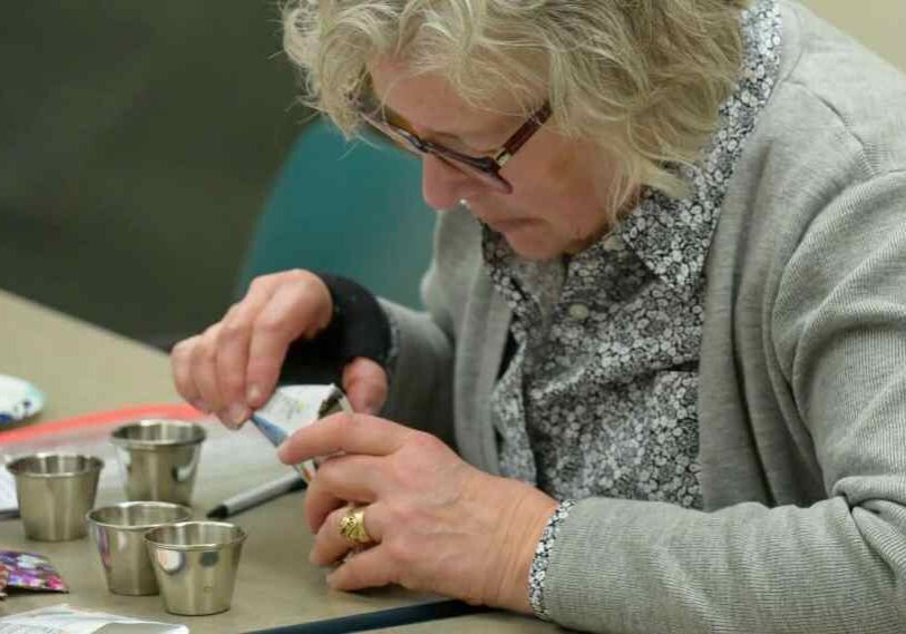 A close-up of an older woman with short, curly gray hair and glasses sitting at a table. She carefully pours seeds from a small packet into one of several small metal cups lined up in front of her. She wears a gray cardigan over a patterned blouse and focuses intently on her task. Papers, seed packets, and a plate sit on the table nearby.