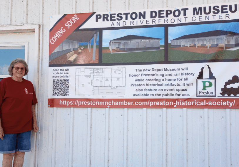 PHOTO 1 Sheila Craig <span style="font-size: 14px; color: #6c6b6b; ">Sheila Craig stands in front of the former Preston Dairy and Farm Building, next to a banner announcing the project to build a Preston Depot Museum and Riverfront Center. (Photo by Alison Leathers)</span>