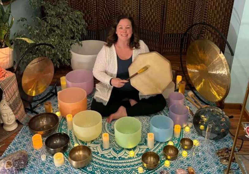 A sound meditation facilitator sits cross-legged on a patterned rug, holding a frame drum and mallet, surrounded by crystal singing bowls, metal bowls, gongs, and softly lit candles in a calm indoor setting.