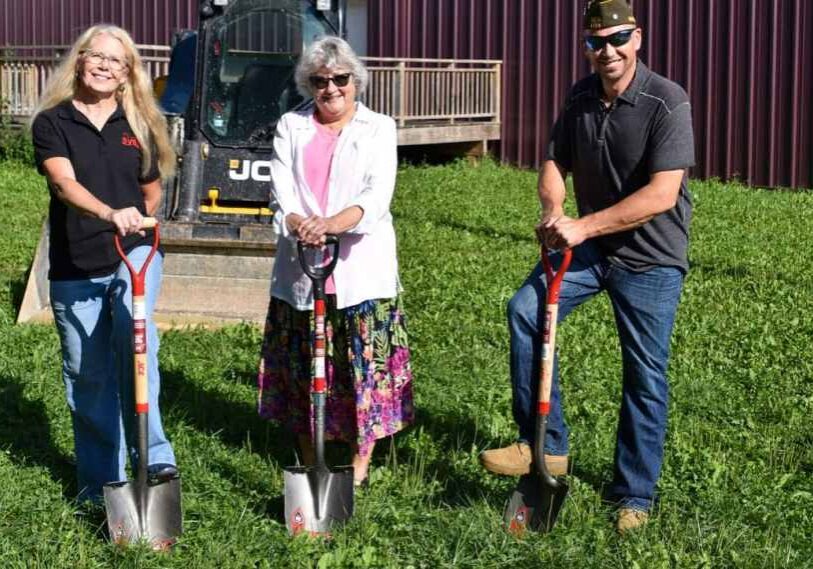 From left to right: two women and a man hold shovels in a grassy area in front of a wooden deck attached to a metal building wall.
