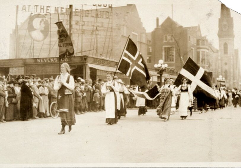 PHOTO 1 Suffage march formatted A black and white photo of women wearing long skirts marching in a parade under various Scandinavian flags.