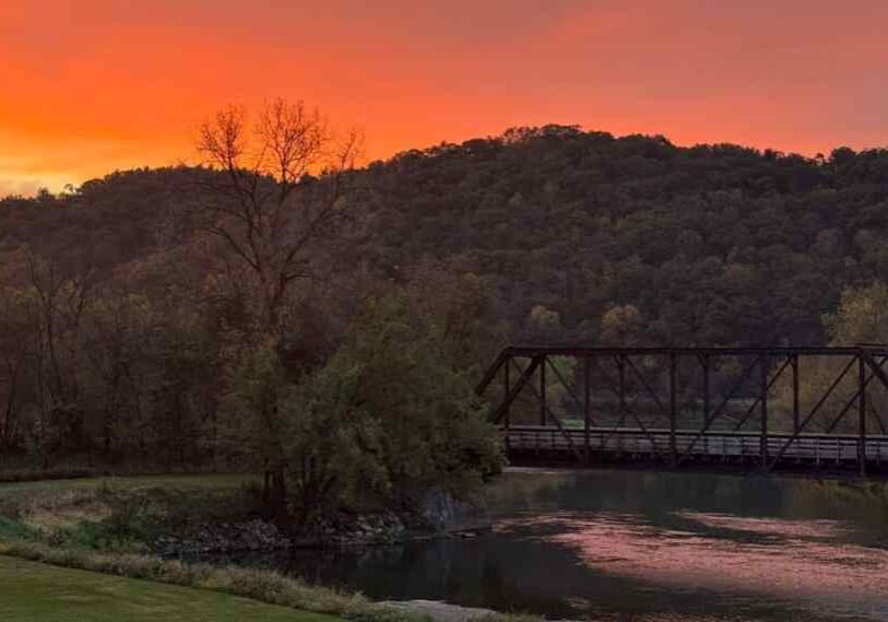PHOTO 1 Sunset over the river Autumn A glowing autumn sunset fills the sky with brilliant shades of orange and red above a wooded bluff. Below, a black steel railroad bridge spans a calm river that reflects the sunset colors. The trees along the water’s edge are beginning to show fall hues of gold and brown, creating a tranquil, scenic view.