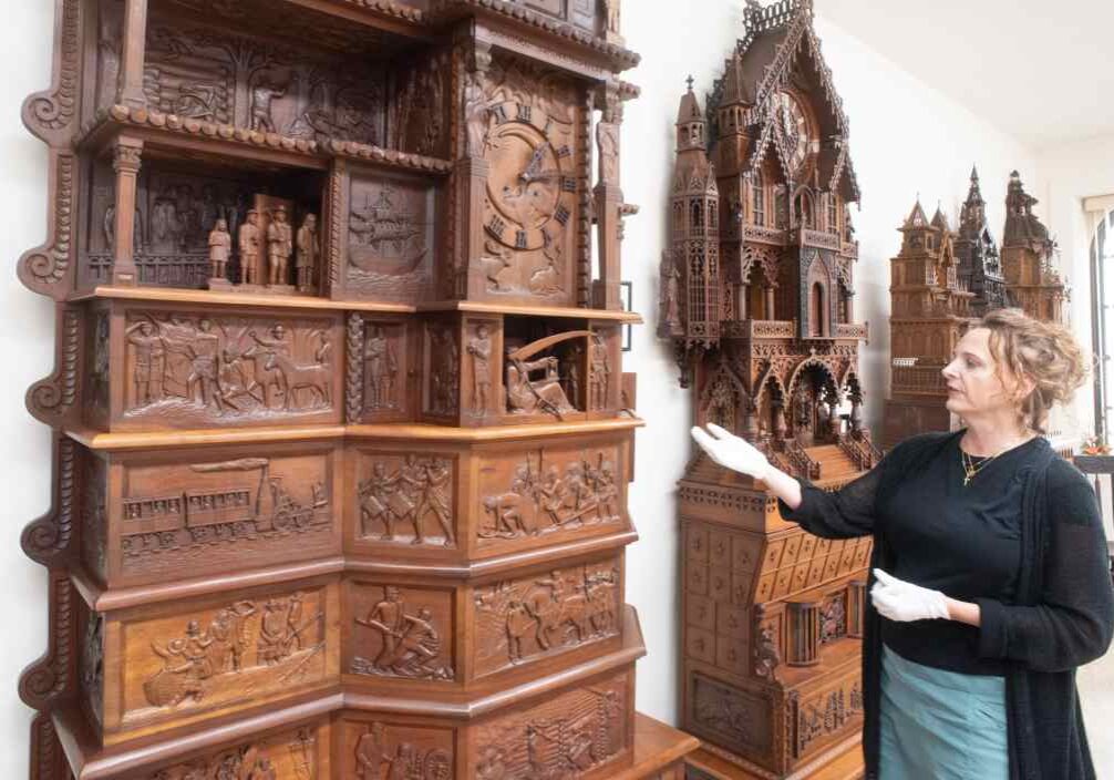 A woman with blonde-brown hair in a loose bun wearing a black sweat set, blue skirt, and white cotton gloves gestures toward an ornately carved 8 foot tall wooden clock.