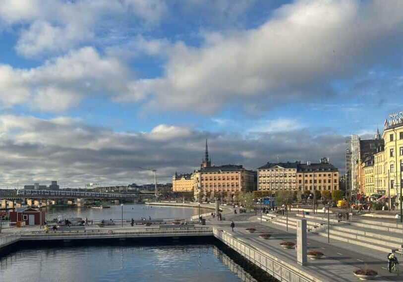 Wide view of Stockholm’s waterfront with bridges, walkways, and historic buildings under a partly cloudy sky, with people walking and biking along the water’s edge.
