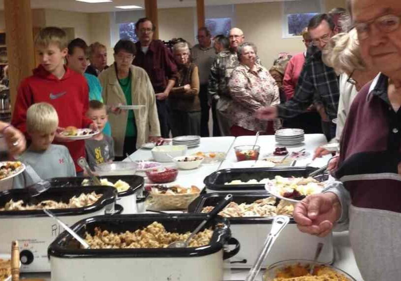 Large group of people of all ages standing in line at a church or community-center buffet, filling their plates with Thanksgiving-style food in large roasters. The room is bright and busy, with casseroles, turkey, salads, and desserts laid out on long tables. The scene shows a warm, friendly small-town gathering.