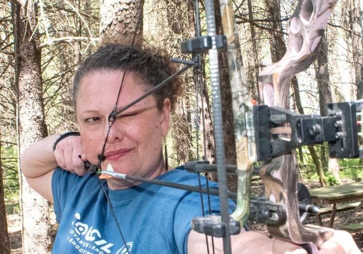 A woman practices archery in a wooded outdoor range, drawing a compound bow and aiming an arrow toward a target. Sunlight filters through tall pine trees as she concentrates on her shot.