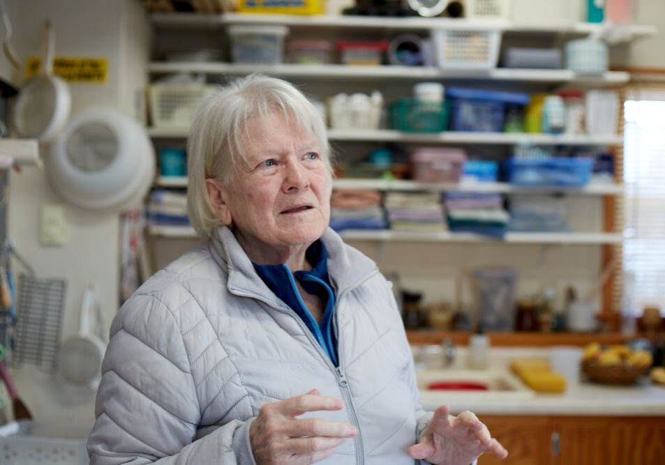 An older woman with short white hair stands in a cozy, well-used workshop or kitchen space, speaking with her hands slightly raised as if explaining something. She wears a light gray quilted jacket over a blue top. Behind her, shelves are filled with containers, tools and supplies, giving the space a creative, hands-on feel. Soft light comes from a window on the right.