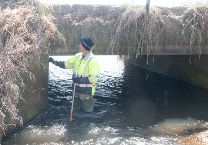 PHOTO-1-The-Bridges-of-Fillmore-County-by-John-Weiss-May-2025 A man in a bright yellow vest stands in a creek under a cement bridge covered in dried brush.