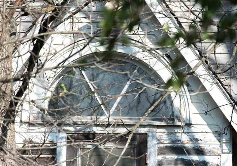 A close-up of an old arched window framed in peeling white paint above a weathered wooden door. Bare tree branches partly obscure the view, and sunlight highlights the cracked glass and faded blue-gray siding.