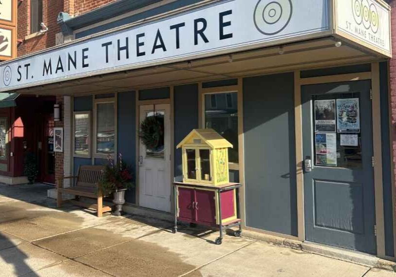 Exterior of the St. Mane Theatre with a vintage marquee on a small-town main street, brick facade, bench and sidewalk in daylight.