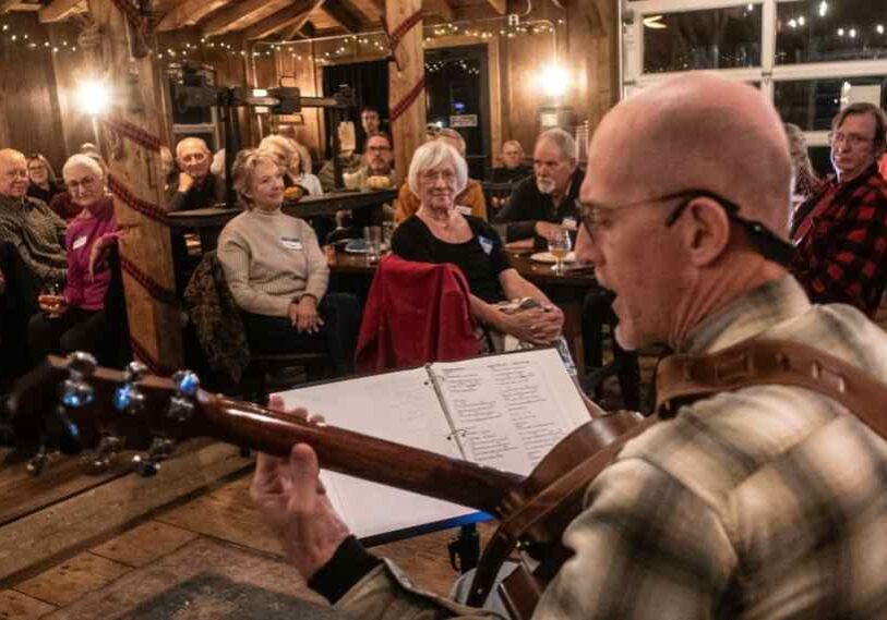A musician wearing glasses and a plaid shirt plays an acoustic guitar on stage inside a warm, wood-paneled venue. He faces an audience of older adults seated at small tables, listening closely under soft string lights.