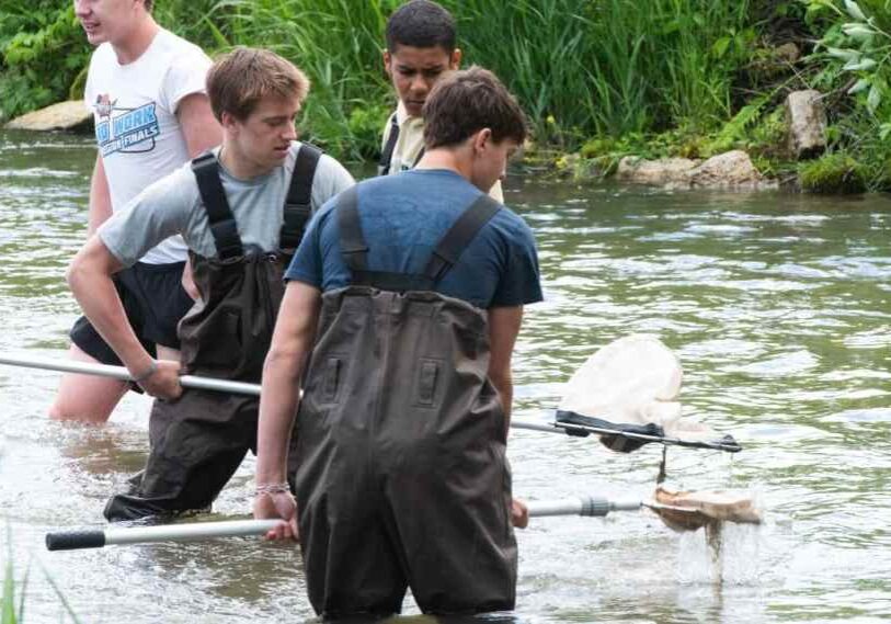 Four high school students, two in chest waders, stand knee deep in a trout stream.