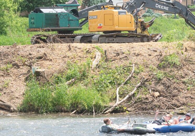 People float down the Root River in tubes while heavy equipment works along the riverbank in the background, where trees and brush have been cleared for a habitat improvement project on a sunny summer day.