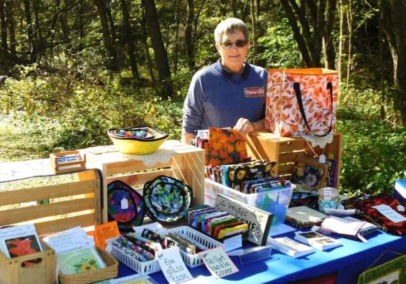 A person with short gray hair and sunglasses stands behind a blue-covered craft fair table set up outdoors on a sunny day in a wooded park. The table displays handmade fabric crafts, including colorful quilted eyeglass cases, pot holders, and bags made from patterned cotton fabric. The vendor’s sign reads “TLT Crafts.” Wooden crates are stacked to create display shelves, and one holds an orange-and-white leaf-patterned tote bag. Dappled sunlight filters through green trees in the background.