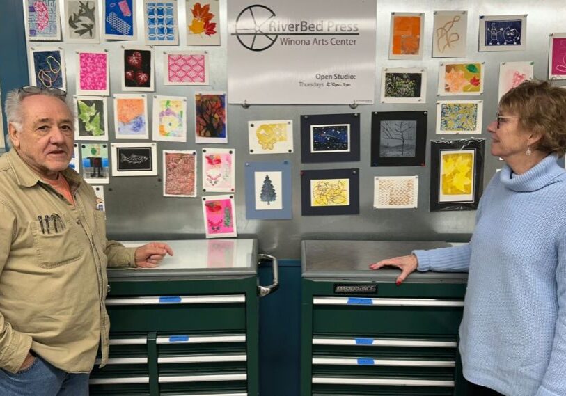 PHOTO 1 Winona Arts Center February 2025 A man in a brown shirt and woman in a light blue sweater stand in front of dark green cabinets and small pieces of art.