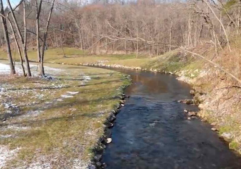 A narrow trout stream curves through a wooded valley with patches of snow on the grassy banks and leafless trees on both sides, showing clear water flowing through a restored creek channel in winter