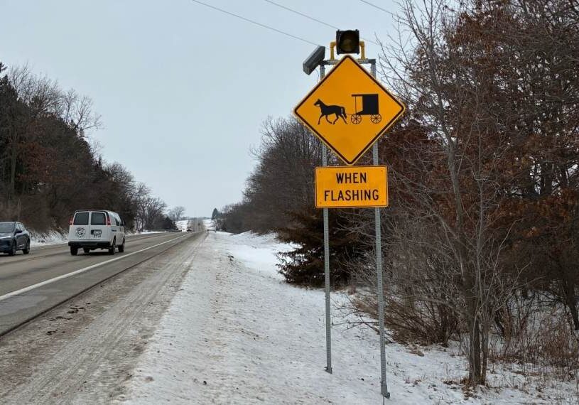 Yellow diamond road sign showing a horse-drawn Amish buggy with a small amber light above it that flashes when a buggy is present, standing beside a snowy rural highway with cars passing on the left and leafless trees lining the roadside in winter