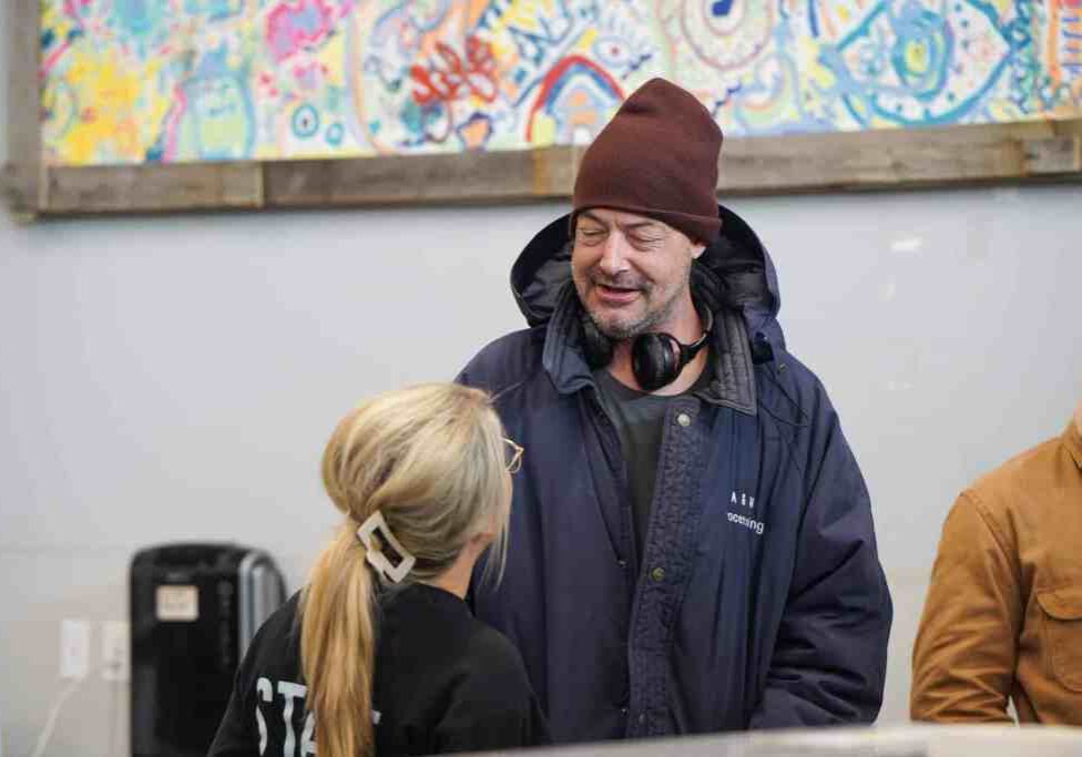 A man wearing a brown knit hat, navy winter coat, and headphones around his neck smiles while talking with a woman whose blonde hair is pulled back with a large cream-colored clip. They stand inside a modern community space with a colorful abstract mural hanging on the wall behind them. Another person in a tan jacket is partially visible at the edge of the photo.