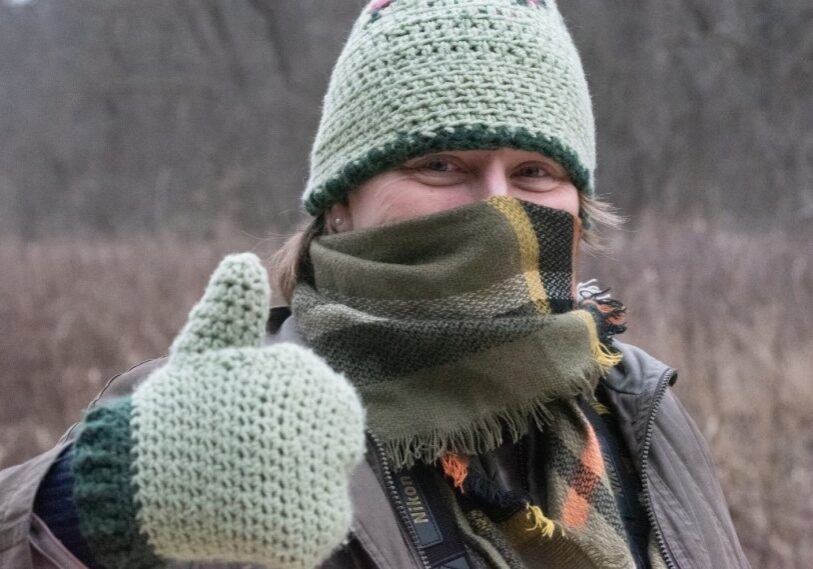 A woman bundled up with a knitted green hat, knitted green mittens, and a plaid scarf gives a thumbs up outside on a winter day.