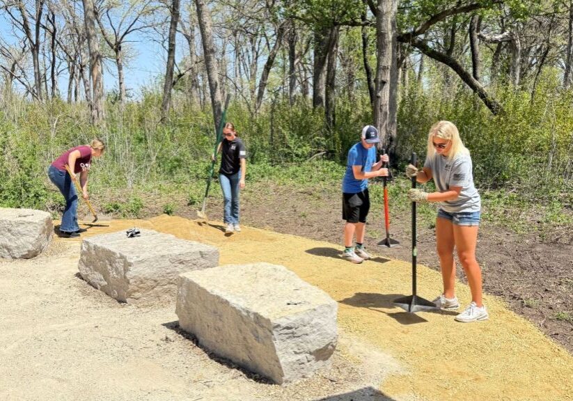Four students hold long metal poles with an x-shaped end that is placed on the gravel ground.