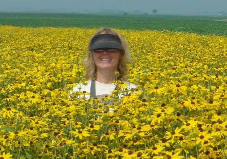 A woman stands in the middle of a wide field filled with bright yellow sunflowers stretching to the horizon. Only her upper body is visible above the flowers. She wears a dark visor and smiles, surrounded by dense blooms under a hazy sky. The landscape is flat and expansive, emphasizing the vast sea of yellow flowers.
