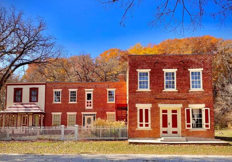 Two red brick historic buildings -- on is two story on the right, the other is one story and stretches out to the left. Windows and doors are trimmed in white wood. Behind the building is a hillside with leaves in yellow and orange against a bright blue sky.