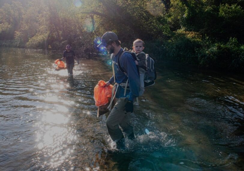 Reid Wintersteen, accompanied by his nine-month old son Clayton, helps with river cleanup on the Root River near Lanesboro. His wife Laura picks up trash nearby. (Photo by John Weiss) 