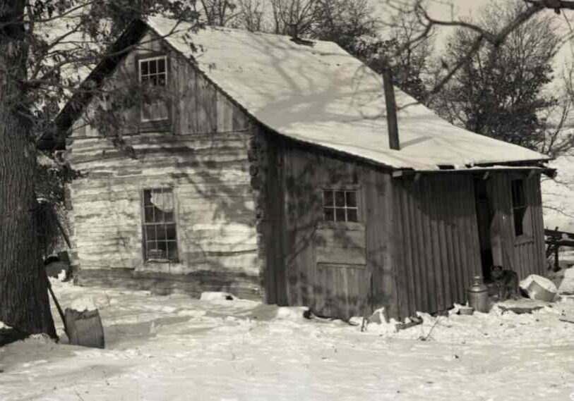 Historic log cabin in winter, partially covered in snow, with weathered wooden walls, small windows and bare trees casting long shadows across a snowy rural yard.