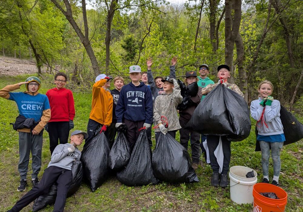 A group of about a dozen children stand together on a wooded trail during a conservation cleanup project in spring. The kids, dressed in hoodies, caps, gloves, and outdoor clothes, proudly hold large black trash bags filled with pulled invasive plants. One child lounges playfully in front while others wave and smile. Buckets and cleanup supplies sit nearby, and fresh green leaves cover the trees and forest floor behind them.