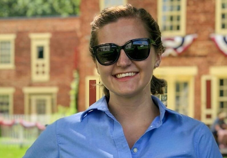 A young woman with light skin smiles while standing outdoors in front of a historic red brick building decorated with patriotic bunting. She wears dark sunglasses and a light blue button-up shirt, with her hair pulled back. The background shows green grass, white fencing, and the restored building, suggesting a historic site setting.