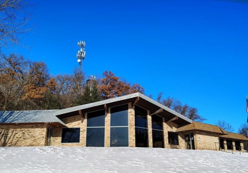 A low, modern brick building sits on a snow-covered slope under a bright blue sky. Large dark windows face outward, reflecting the light. Bare trees line the hillside behind the building, and a tall communications tower rises above the trees. An American flag stands on a pole near the right side of the building.