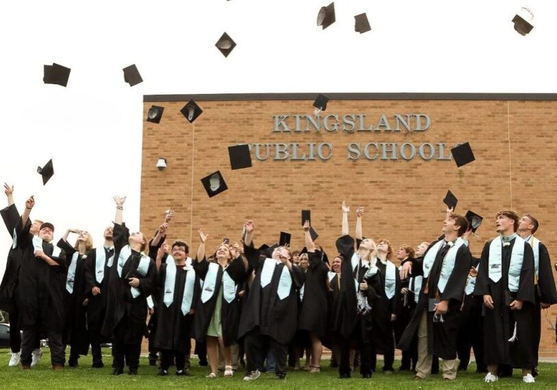 Students in black graduation gowns stand in front of an orange brick building and throw their black caps into the air.