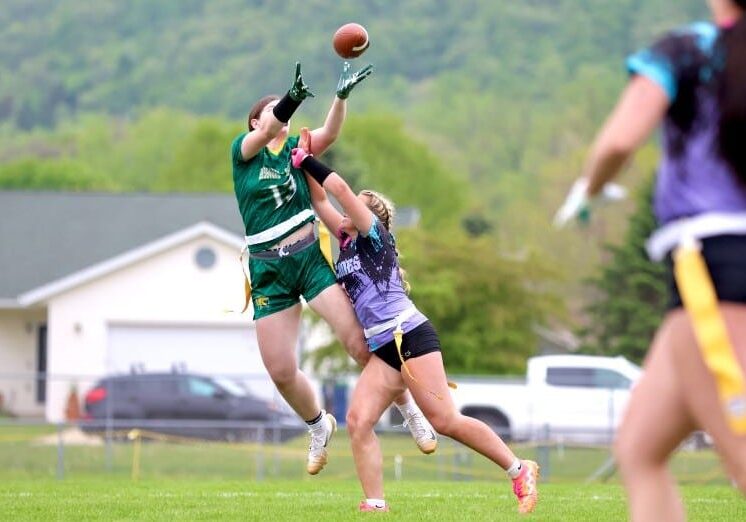 Two high school girls leap into the air while competing for a football during a flag football game. Players in colorful uniforms watch the contested catch from the field.