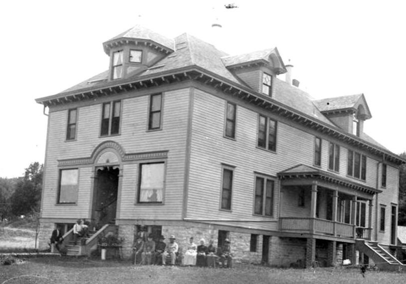 Black and white photo of an old two-story rectangular building with windows wrapping around the building.