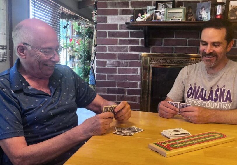 RRC Neighbors in Action February 2025 Two men, one older and one younger, play cribbage on a wood table in front of a red brick fireplace.