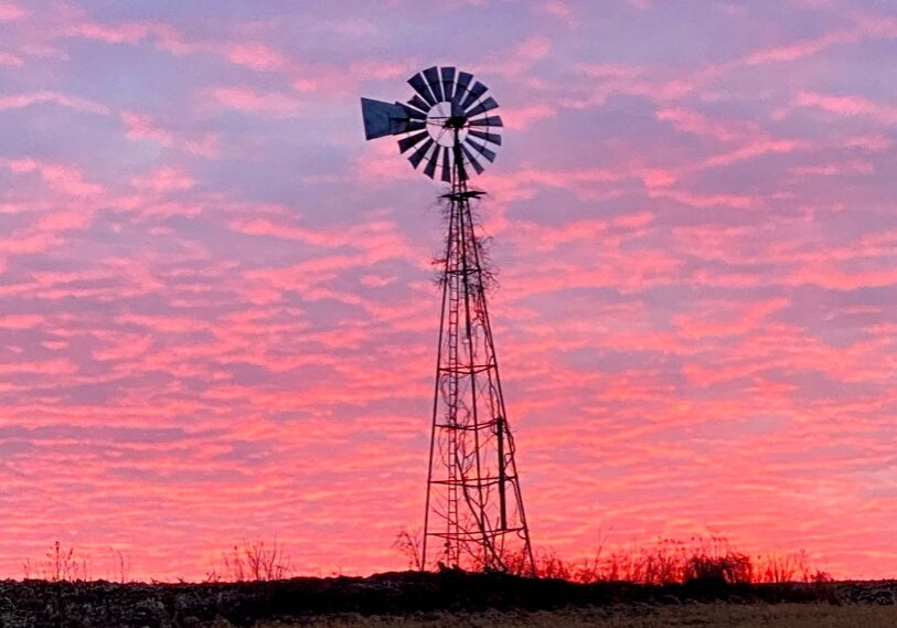 An old fashioned metal windmill stands above a field against a sky of pink and blue clouds at sunset