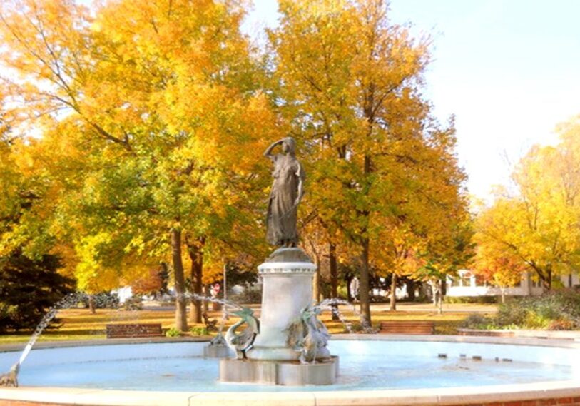 A stone statue of a woman stands at the center of a circular fountain, with water spraying outward from sculpted fish at the base. The statue is surrounded by trees with bright yellow and orange autumn leaves. Benches and a lawn are visible in the background, and sunlight illuminates the park setting.